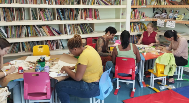 #paratodosverem: na imagem, há uma biblioteca com estantes bege repletas de livros de diversas cores e com algumas bonecas. Em foco, estão duas mesas coloridas, que apoiam as sacolas. Sobre as mesas, também há papéis, pincéis e tintas de diversas cores. Ao redor das mesas, sentadas em cadeiras coloridas, funcionárias da escola em trajes diversos pintam sacolas. 