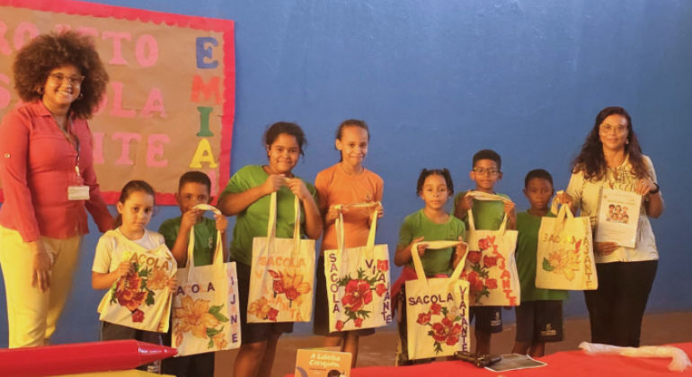 #paratodosverem: em um espaço coberto, sete crianças uniformizadas estão lado a lado, segurando sacola decorada com desenhos de flores na qual está escrito “Sacola Viajante”. Ao lado delas, duas mulheres adultas, uma de camisa vermelha e outra de blusa estampada, também posam para a foto. Ao fundo, há uma parede azul em que está fixado um mural colorido com a frase “Projeto Sacola Viajante”.