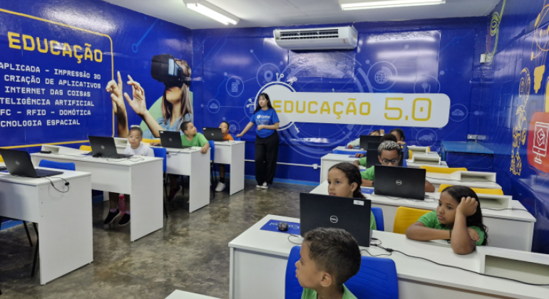 #paratodosverem: fotografia de sala de aula com paredes azuis em que está escrito “Educação 5.0”. Há várias crianças uniformizadas sentadas, sozinhas ou em duplas, mexendo em computadores, com uma educadora auxiliando-as.