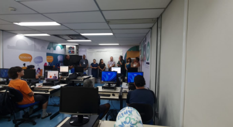 #paratodosverem: em uma sala equipada com computadores, seis estudantes estão sentados(as) em suas mesas, atentos(as) a oito formadores(as) que estão em pé, sobre um piso azul, à frente da sala. Atrás destes(as), está afixado um quadro branco, e, nas paredes laterais, há uma decoração colorida com palavras como “tecnologia” e “cidadão”. 