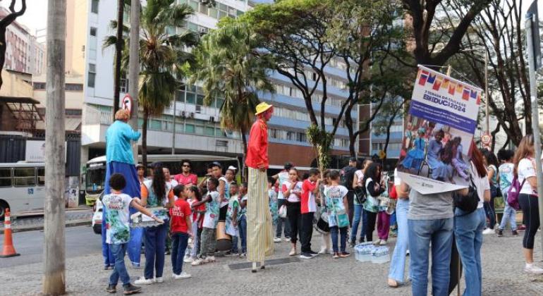 #paratodosverem: A imagem mostra uma movimentada atividade de rua da “3ª Semana da Educação BH EDUCA 2024”. Crianças com camisetas coloridas e adultos participam do evento. Dois artistas em pernas de pau, fantasiados, interagem com o público. Ao fundo, há prédios altos e árvores. À direita, uma pessoa segura um cartaz do evento. O clima é de celebração e alegria.