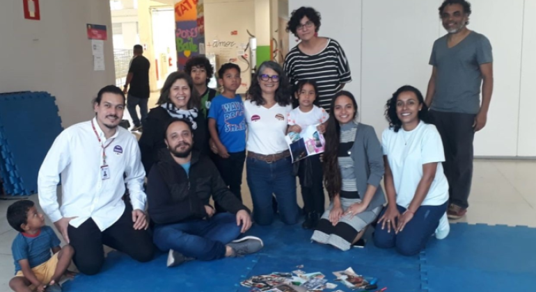 #paratodosverem: um grupo de pessoas adultas e crianças posa para foto, sobre um tatame azul, em um espaço interno. Sobre o tatame, há folhas de papel, fotos, tesouras e caixas de lápis. Algumas pessoas estão sentadas, outras de pé. Ao fundo, paredes brancas e colunas, com cartazes coloridos.