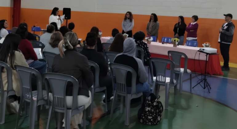 #paratodosverem: na imagem, pessoas sentadas em cadeiras cinzas acompanham uma apresentação, em um ginásio com paredes pintadas nas cores laranja e branco. À frente, seis pessoas estão em pé, próximas a uma mesa com toalha rosa e vasos de flores, e uma mulher segura o microfone. No canto direito, um projetor sobre tripé aponta para a parede.