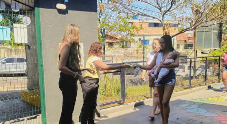 #paratodosverem: Uma mulher de blusa amarela e calça preta entrega um panfleto a uma jovem de camiseta branca sem mangas e shorts jeans, que segura um bebê no colo. Ao lado, outra mulher de preto observa. Atrás, há uma senhora de blusa branca e saia estampada. A cena ocorre em área externa ensolarada, com muros, árvores e casas ao fundo.