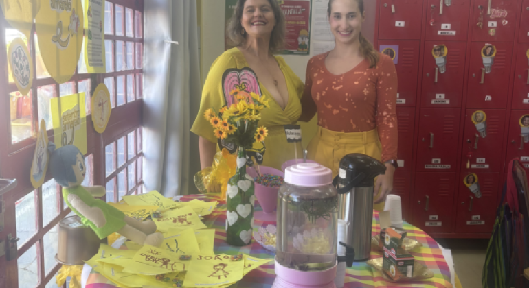 #paratodosverem: Duas mulheres sorriem atrás de uma mesa decorada com toalha colorida, flores amarelas, potes de doces e recipientes de bebidas. Sobre a mesa há também desenhos infantis em folhas amarelas e um boneco de pelúcia azul e verde. Ao fundo, há armários vermelhos com fotos e nomes. Na parede e na janela aparecem cartazes com o tema “Setembro Amarelo”.