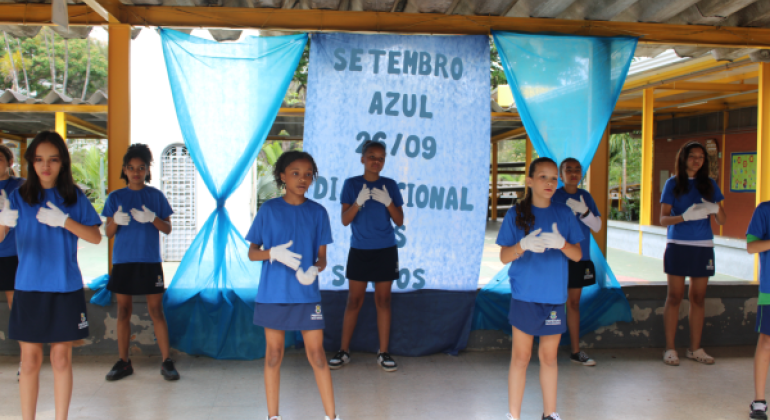 #paratodosverem: A imagem mostra um grupo de crianças se apresentando em frente a um painel azul com os dizeres “Setembro Azul 26/09 Dia Nacional dos Surdos”. Elas vestem camisetas azuis, shorts ou saias da mesma cor e usam luvas brancas, posicionadas em gestos que sugerem linguagem de sinais. Ao fundo, há cortinas azuis presas a uma estrutura amarela.