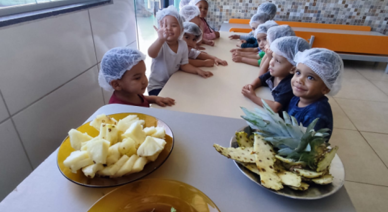 #paratodosverem: A imagem mostra um grupo de crianças sentadas em volta de uma mesa, todas usando toucas higiênicas brancas. À frente, há pratos com pedaços de abacaxi, cascas da fruta e folhas verdes, possivelmente hortelã. As crianças parecem atentas e sorridentes, participando de uma atividade em um ambiente escolar com paredes de azulejo colorido.