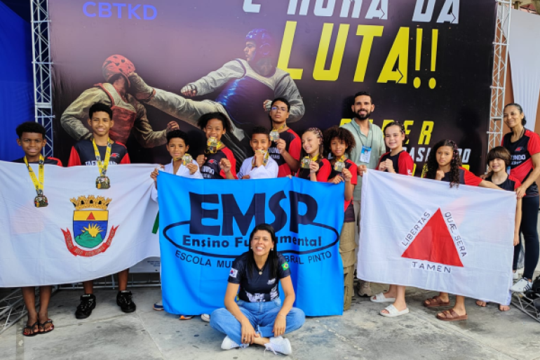 #paratodosverem: Em um evento de taekwondo, um grupo de jovens atletas e instrutores está posando com orgulho em frente a um banner. As crianças exibem medalhas no peito e seguram três bandeiras: o brasão municipal, uma faixa azul da escola ("EMSP") e a bandeira de Minas Gerais. 