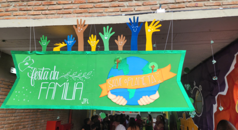 #paratodosverem: na foto, há um painel verde, em primeiro plano, com moldura brilhante, que exibe a frase “Festa da Família”, a mensagem “Salve o Planeta” sobre um desenho da Terra e mãos segurando-a. Acima, mãos coloridas nas cores verde, azul, marrom e amarelo, com rostos sorridentes, completam a decoração. Ao fundo, pessoas conversam em um corredor decorado com murais.