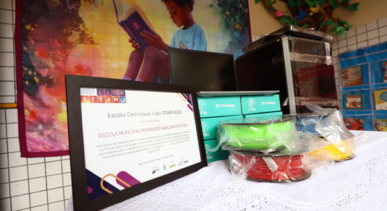 Escola recebe o kit de impressora 3D como destaque da Liga STEAM 2025. #paratodosverem: A imagem mostra uma mesa coberta com toalha branca, onde estão dispostos um certificado de destaque da Liga STEAM e caixas com materiais coloridos de filamentos para impressora 3D. Ao fundo, há um painel ilustrado com a imagem de uma criança lendo um livro.