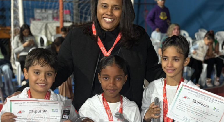 #paratodosverem: em um ginásio esportivo, uma mulher vestida de preto sorri enquanto abraça três crianças que seguram diplomas e medalhas. As crianças vestem uniformes brancos de taekwondo e usam medalhas vermelhas no pescoço. A mulher também usa uma medalha semelhante. Ao fundo, há cadeiras brancas ocupadas por outras pessoas e uma quadra com rede de futsal. O clima é de comemoração.