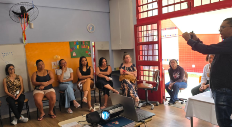#paratodosverem: a foto mostra uma roda de conversa em uma sala de aula, onde um grupo de pais e responsáveis assiste à apresentação de Telly Almeida. Ele está em pé, gesticulando. Um projetor está ligado sobre uma mesa, ao lado de um notebook, no centro da sala. Ao fundo, há um cartaz com a frase "Feliz Aniversário" e desenhos infantis afixados em uma parede laranja.
