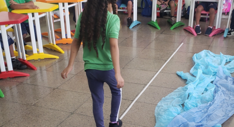 #paratodosverem: Em sala de aula, uma menina caminha sobre uma linha branca no chão de uma sala, enquanto outras crianças, sentadas em carteiras coloridas, observam. Ao lado da linha, há um tecido azul amontoado no chão. Ao fundo, vêem-se mochilas penduradas na parede