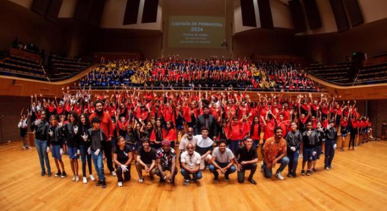 #paratodosverem: Foto tirada em auditório amplo e moderno, com palco e plateia. No centro, centenas de crianças uniformizadas com camisetas coloridas (vermelhas, azuis, amarelas e pretas) formam um grande coral escolar. À frente, adultos e jovens, possivelmente educadores e artistas, posam sorrindo. Ao fundo, projeção: “Cantata de Primavera 2024”.