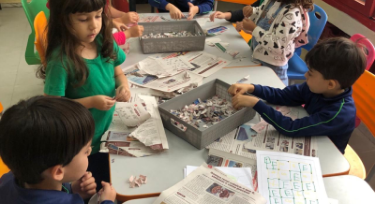 #paratodosverem: seis crianças estão sentadas e em pé ao redor de uma mesa, rasgando folhas de jornal em pedaços pequenos. No centro, há duas bandejas com tiras e pedaços de papel. A sala é clara, com paredes amarelas e janelas de moldura vermelha. Sobre a mesa também há folhas com desenhos e atividades escolares coloridas.