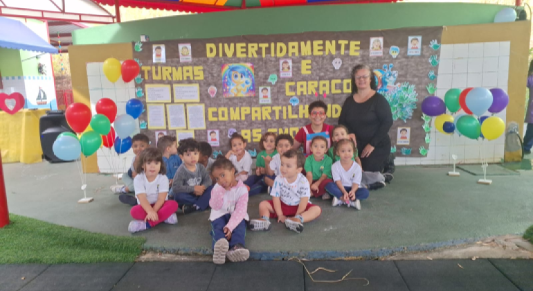 #paratodosverem: A foto mostra um grupo de crianças sentadas no chão em frente a um mural decorado, acompanhadas por uma mulher em pé ao lado direito. O mural tem o tema “Divertidamente e Caracol Compartilhando as Emoções”, com letras grandes em amarelo, desenhos coloridos, mãos pintadas e fotos das crianças coladas. Em volta, há balões coloridos em suportes brancos. A cena acontece sob uma cobertura de lona colorida.