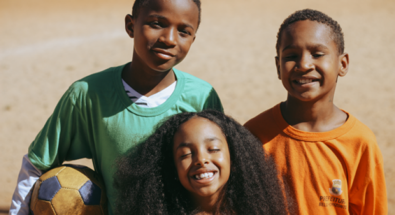 #paratodosverem: Em um close-up, duas crianças, um menino de uniforme verde e um de uniforme laranja, olham sorrindo para a câmera. Entre eles, em primeiro plano, uma menina com cabelos cacheados e volumosos sorri de olhos fechados. O menino de verde segura uma bola de futebol na lateral. O sol ilumina os rostos do grupo, transmitindo alegria e conexão.