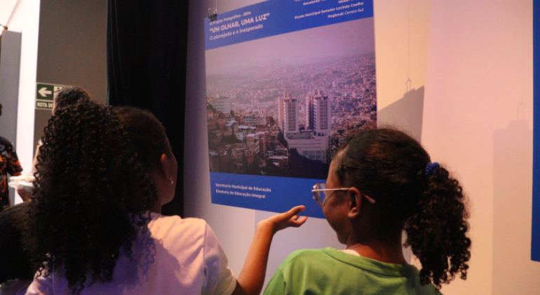 #paratodosverem: duas crianças observam atentamente uma fotografia exposta em um painel azul, parte do projeto “Um Olhar, Uma Luz – 2024”. A imagem mostra uma vista urbana de Belo Horizonte. O ambiente está com luz suave, e o momento transmite curiosidade e interesse das crianças pela arte fotográfica.