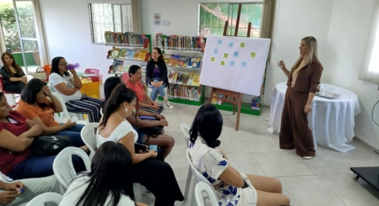 #paratodosverem: a imagem mostra uma sala pintada de branco, com estantes repletas de livros entre duas janelas gradeadas ao fundo. Sentadas em cadeiras brancas de plástico, pessoas adultas estão atentas a uma mulher em pé, ao lado de um cavalete que sustenta um quadro com post-its verdes e azuis. Atrás da palestrante, há uma mesa redonda coberta por toalha branca.
