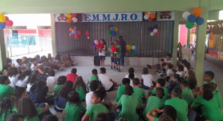#paratodosverem: a imagem mostra um grupo de estudantes reunidos(as) em espaço coberto da EM Monsenhor João Rodrigues de Oliveira. Três crianças, com fantoches, apresentam-se à frente dos(as) colegas, acompanhadas por uma professora. Balões coloridos decoram o local. Os(As) demais alunos(as) assistem à apresentação sentados(as).