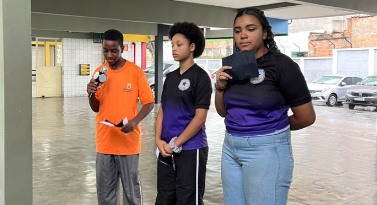 #paratodosverem: em uma área semiaberta da escola, três estudantes estão de pé, um garoto, à esquerda, e duas garotas, à direita. O menino segura um microfone enquanto lê seu discurso em um papel que segura, enquanto as meninas aguardam o momento de falar.
