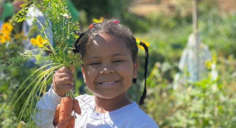 #paratodosverem: A imagem mostra uma criança sorridente segurando cenouras recém-colhidas com as folhas ainda presas, em um ambiente de horta. Ela veste uma camiseta branca com o brasão e a inscrição "Prefeitura de Belo Horizonte". Ao fundo, há vegetação verde e flores amarelas.
