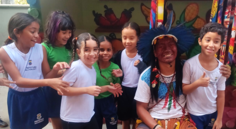 #paratodosverem: na foto, um indígena Pataxó, com cocar azul, colares e pintura facial, sorri ao lado de crianças uniformizadas. Elas também estão sorrindo. Ao fundo, há uma parede com pinturas de alimentos e um tecido com rosto indígena colorido, além de pássaros e flores.