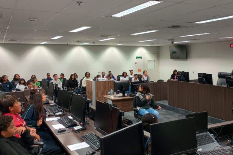 #paratodosverem: A imagem mostra uma sala ampla com mesas em formato de “U” equipadas com computadores e microfones. Diversas crianças e adolescentes estão sentados nas cadeiras, alguns na parte central e outros nas laterais, enquanto uma menina segura um microfone. Ao fundo, há mais pessoas sentadas em fileiras. O ambiente é um plenário moderno.
