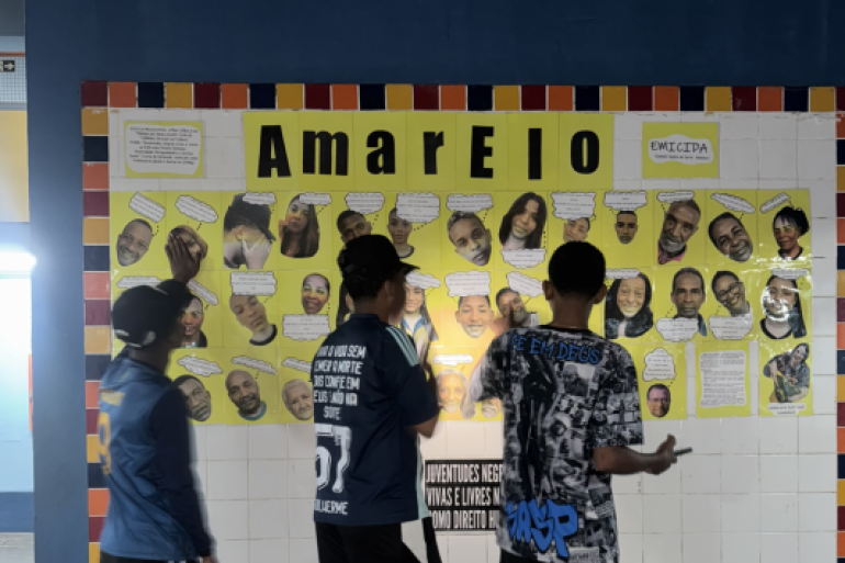A música encorajou os estudantes a compartilhar experiências #paratodosverem: Três jovens estão de frente para um grande mural de azulejos, com o título "Amarelo" em destaque. O mural exibe recortes de cabeças de várias pessoas sorrindo e textos inspirados no álbum de Emicida. Os jovens, de costas, estão concentrados, lendo o conteúdo e interagindo com a arte em um corredor escolar.