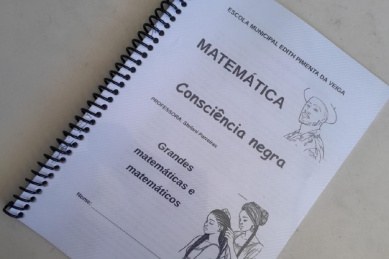 As apostilas retratam figuras negras importantes para a matemática. #paratodosverem: Capa de uma apostila com encadernação em espiral preta. O título é "Matemática Consciência negra" e o nome da professora é "Stefani Parreiras". O material inclui ilustrações em preto e branco de figuras afro-brasileiras e espaços para o nome do aluno e turma. O topo da capa tem o nome da escola: "ESCOLA MUNICIPAL EDITH PIMENTA DA VEIGA".