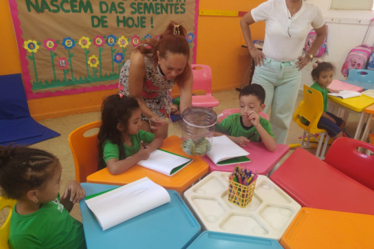 #paratodosverem: A imagem mostra uma sala de aula colorida com mesas e cadeiras infantis. Quatro crianças com camisetas verdes estão sentadas desenhando em cadernos, enquanto uma professora mostra as borboletas dentro de um pote transparente. Outra mulher observa em pé. Ao fundo, há um mural com flores e a frase “Nascem das sementes de hoje!”