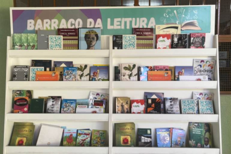 #paratodosverem: A imagem mostra uma estante branca com várias prateleiras repletas de livros de diferentes tamanhos e cores. Na parte superior há uma faixa com o título “Barraco da Leitura” em letras grandes brancas sobre um fundo colorido. Entre os livros, é possível ver obras literárias infantis e juvenis, além de alguns títulos voltados à arte e cultura afro-brasileira. O ambiente é iluminado e tem uma parede verde clara ao fundo.