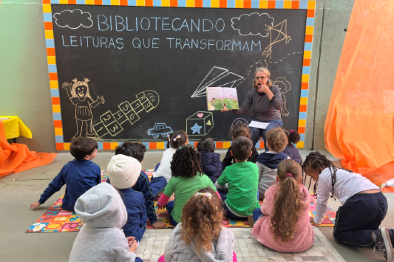 #paratodosverem: Em um espaço escolar decorado com um grande quadro preto, uma professora de blusa cinza lê um livro para um grupo de crianças pequenas, sentadas no chão. O quadro exibe a frase "BIBLIOTECANDO - LEITURAS QUE TRANSFORMAM". A cena, de fundo laranja vibrante, transmite um momento de contação de histórias e incentivo à leitura.