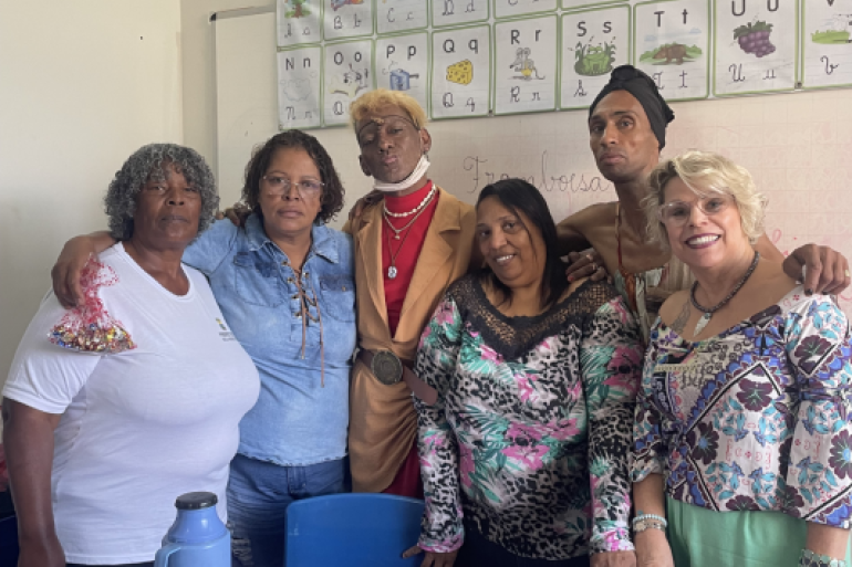 #paratodosverem: Um grupo de seis pessoas, a maioria professoras e funcionárias, posa abraçado em uma sala de aula. A cena, em frente a um quadro com o alfabeto, sugere um momento de confraternização e celebração pedagógica, com um toque criativo e festivo.