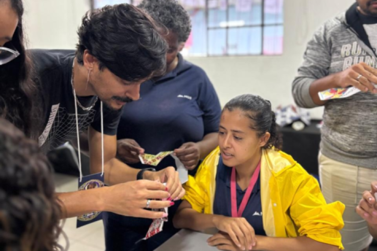 O projeto visou a integrar os ensinos de geometria e artes. #paratodosverem: Duas mulheres e dois homens adultos, além de uma estudante de jaqueta amarela, estão reunidos em torno de uma mesa, em um close-up. O grupo examina pequenos pedaços de papel. A estudante de jaqueta amarela e um dos homens olham atentamente para os papéis nas mãos. O ambiente é um espaço interno.