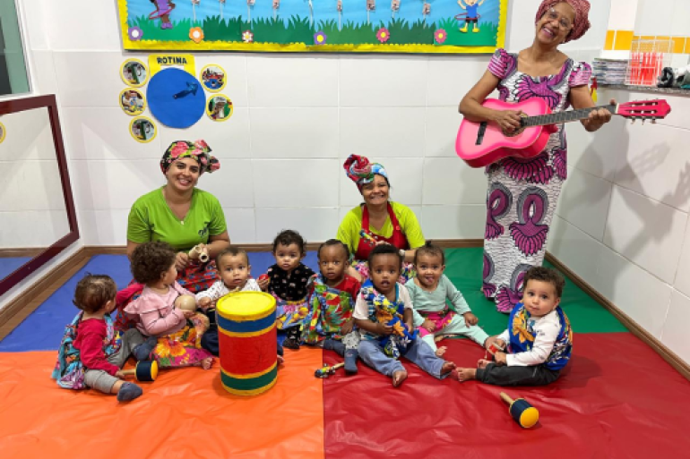 #paratodosverem: A imagem mostra uma sala iluminada e bebês sentados em um tapete colorido, segurando instrumentos musicais como chocalhos e tambores, acompanhados por três educadoras que usam roupas e turbantes coloridos. Uma delas está em pé tocando um violão rosa, as outras estão sentadas. Ao fundo, há um mural decorado com personagens infantis.