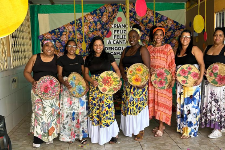 #paratodosverem: A imagem mostra oito mulheres sorridentes em uma apresentação cultural. Elas vestem saias longas coloridas e seguram peneiras decoradas com flores. Ao fundo, há um painel com tecidos estampados