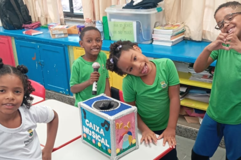 #paratodosverem: Quatro crianças estão em uma sala de aula, sorrindo em volta de uma caixa colorida com o nome “Caixa Musical”. Duas delas vestem camisetas verdes e duas, brancas, todas com o logotipo da Prefeitura de Belo Horizonte. O ambiente é alegre, com mesas, cadeiras vermelhas e prateleiras azuis cheias de livros e materiais escolares.