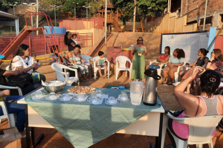 Pais e responsáveis participam de rodas de conversa na escola #paratodosverem: A imagem mostra um encontro de pais ao ar livre, com participantes sentados em círculo enquanto uma mulher conduz a conversa. Há uma mesa central com lanche, copos e uma garrafa térmica.