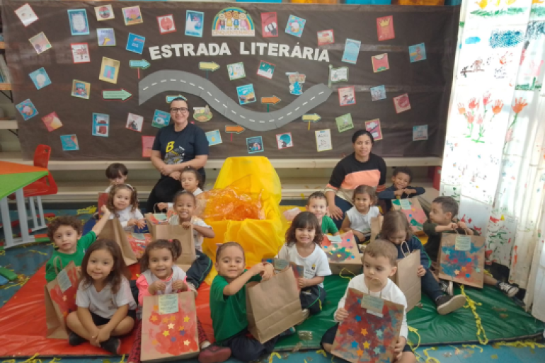 Projeto visitou cidades mineiras #paratodosverem: Em uma sala decorada com a frase "Estrada Literária", um grupo de crianças pequenas está sentado no chão com suas professoras. As crianças seguram sacolas de papel pardo decoradas com estrelas coloridas. O cenário é festivo, com confetes e um fundo que simula uma estrada com livros.