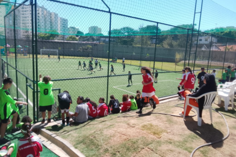 Estudantes se divertiram no festival esportivo “Joga Integrada” #paratodosverem: Crianças assistem e participam de um jogo de futebol em um campo cercado por tela. Alguns jogadores estão em campo, outros, de uniforme vermelho ou verde, aguardam do lado de fora. Há pessoas sentadas em cadeiras plásticas brancas, enquanto prédios e árvores aparecem ao fundo sob um céu azul.