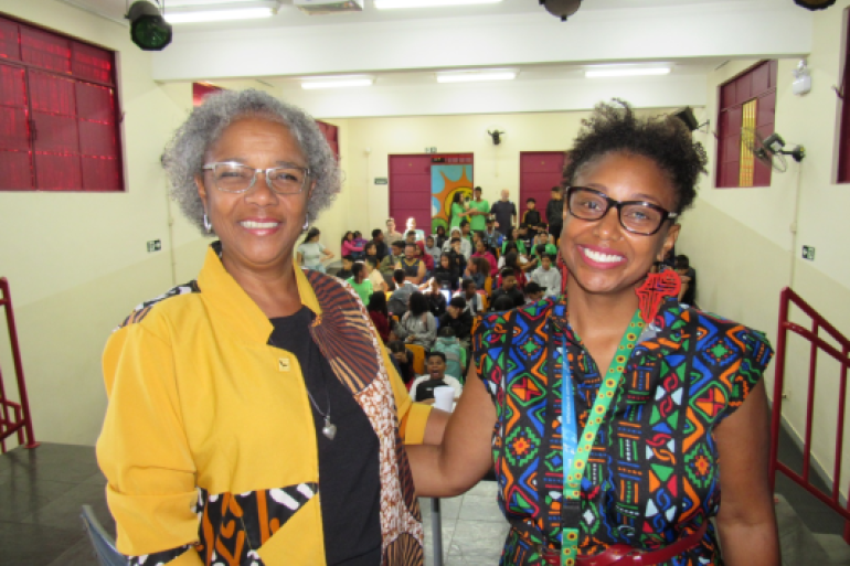 Estudantes participaram de encontro com professora doutora da UFMG #paratodosverem: Duas mulheres sorriem para a câmera em auditório cheio de estudantes ao fundo. Ambas usam roupas coloridas com estampas afro-brasileiras. A mulher à esquerda tem cabelos grisalhos curtos e veste uma blusa amarela com detalhes estampados; a mulher à direita usa óculos, brincos grandes vermelhos e um vestido estampado.