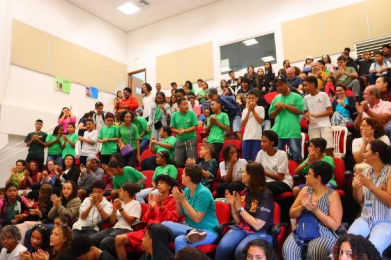 #paratodosverem: Interior de um auditório com degraus e poltronas vermelhas ocupadas por um grande grupo de jovens e adultos. Muitos estudantes vestem camisetas verdes de uniforme e aparecem aplaudindo. As paredes são brancas com painéis retangulares claros. A iluminação vem de luminárias quadradas no teto.