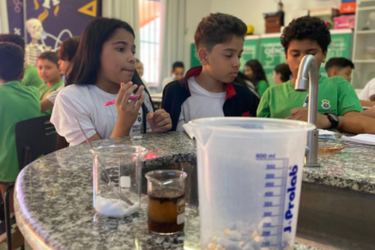#paratodosverem: A imagem mostra estudantes participando de uma atividade científica em um laboratório escolar. Eles observam atentamente materiais como béqueres e um copo medidor sobre a bancada de granito, enquanto discutem e fazem anotações. 
