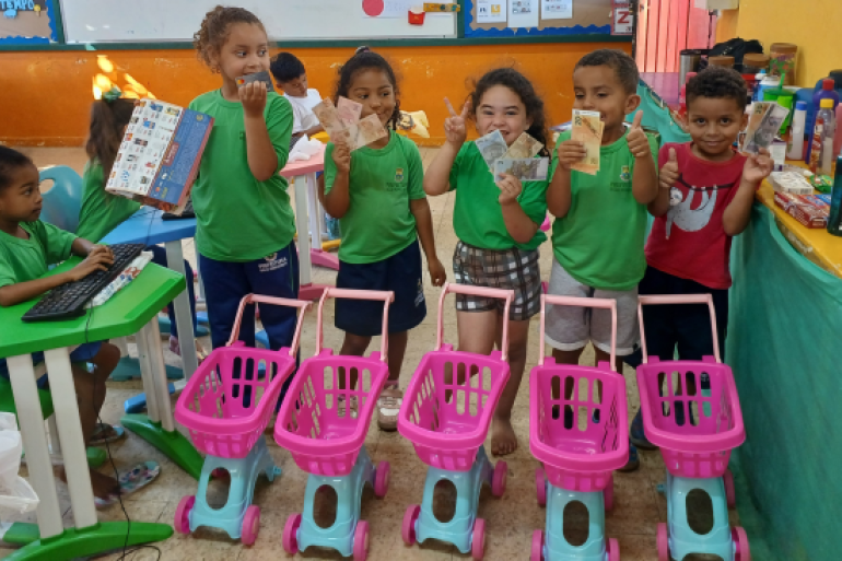 #paratodosverem: A imagem mostra sete crianças em uma sala de aula colorida. Seis delas estão em pé, sorrindo e segurando notas de dinheiro de brinquedo, e uma está sentada digitando em um teclado. À frente delas há quatro carrinhos de compras de brinquedo rosa e azul. Ao fundo, sobre uma mesa coberta por um tecido verde, há diversos produtos, como xampus, sabonetes e cremes, simulando um mercado. As crianças vestem uniformes verdes da rede municipal e demonstram alegria durante a atividade.