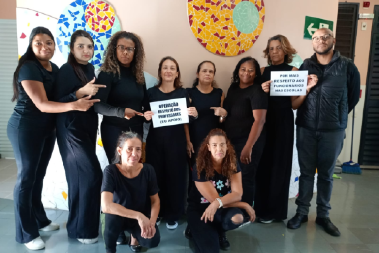 #paratodosverem: Um grupo de dez funcionários escolares, em sua maioria mulheres, está posando em frente a uma parede decorada com mosaicos. Eles vestem roupas pretas e seguram cartazes com mensagens de protesto, como "Operação Respeito aos Professores (Eu Apoio)”.