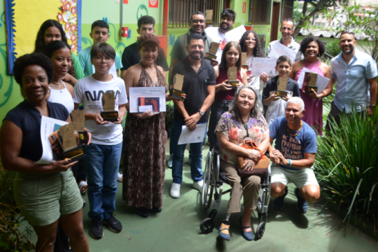 #paratodosverem: Um grupo misto de adultos e jovens, cerca de 16 pessoas, posa em pé e ajoelhado em um pátio externo da escola, cercado por folhagens. Vários membros do grupo seguram troféus, certificados ou pequenos objetos de premiação. Uma mulher está sentada em uma cadeira de rodas no centro. A parede atrás é pintada em cores vivas, incluindo verde e amarelo.