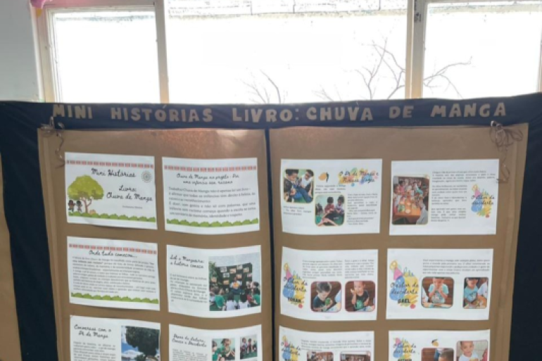 #paratodosverem: Um painel de exposição de papel pardo e preto com o título "MINI HISTÓRIAS LIVRO: CHUVA DE MANGA" está fixado em frente a uma janela. O painel exibe vários cartões de texto e fotos das crianças, organizados em colunas. As janelas, ao fundo, mostram uma paisagem externa com galhos de árvores secas.