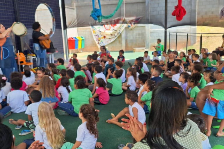 #paratodosverem: A imagem mostra um grupo grande de crianças sentadas no chão, assistindo a uma apresentação musical. Duas pessoas tocam instrumentos, uma com pandeiro e outra com violão, diante do público infantil. O evento ocorre em um espaço coberto, com parede azul e brinquedos coloridos ao fundo.