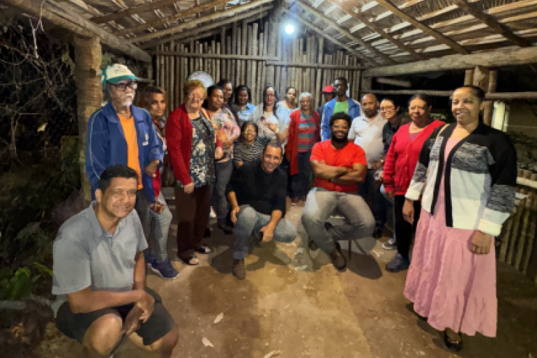 #paratodosverem: Um grupo de adultos posa em uma estrutura rústica de madeira e palha,. As pessoas aparecem sorrindo e reunidas.